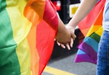 População LGBTQIA+ pode usar ata notarial para denunciar ataques virtuais Casal LGBTQIA+ de mãos dadas com a bandeira do arco iris